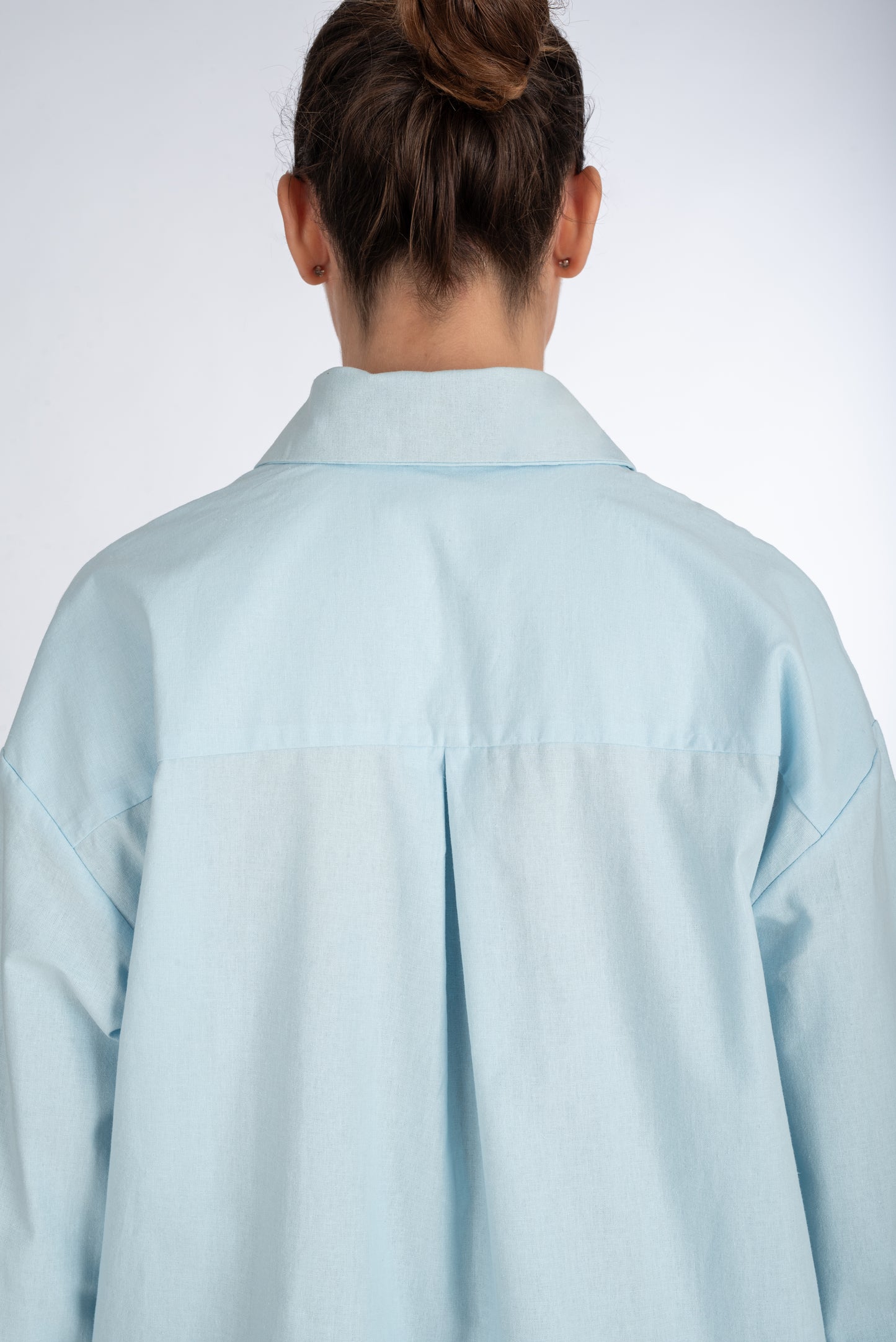 Lifestyle shot of the back of a baby blue women's shirt, highlighting the center pleat at the yoke and sleek, minimal design.