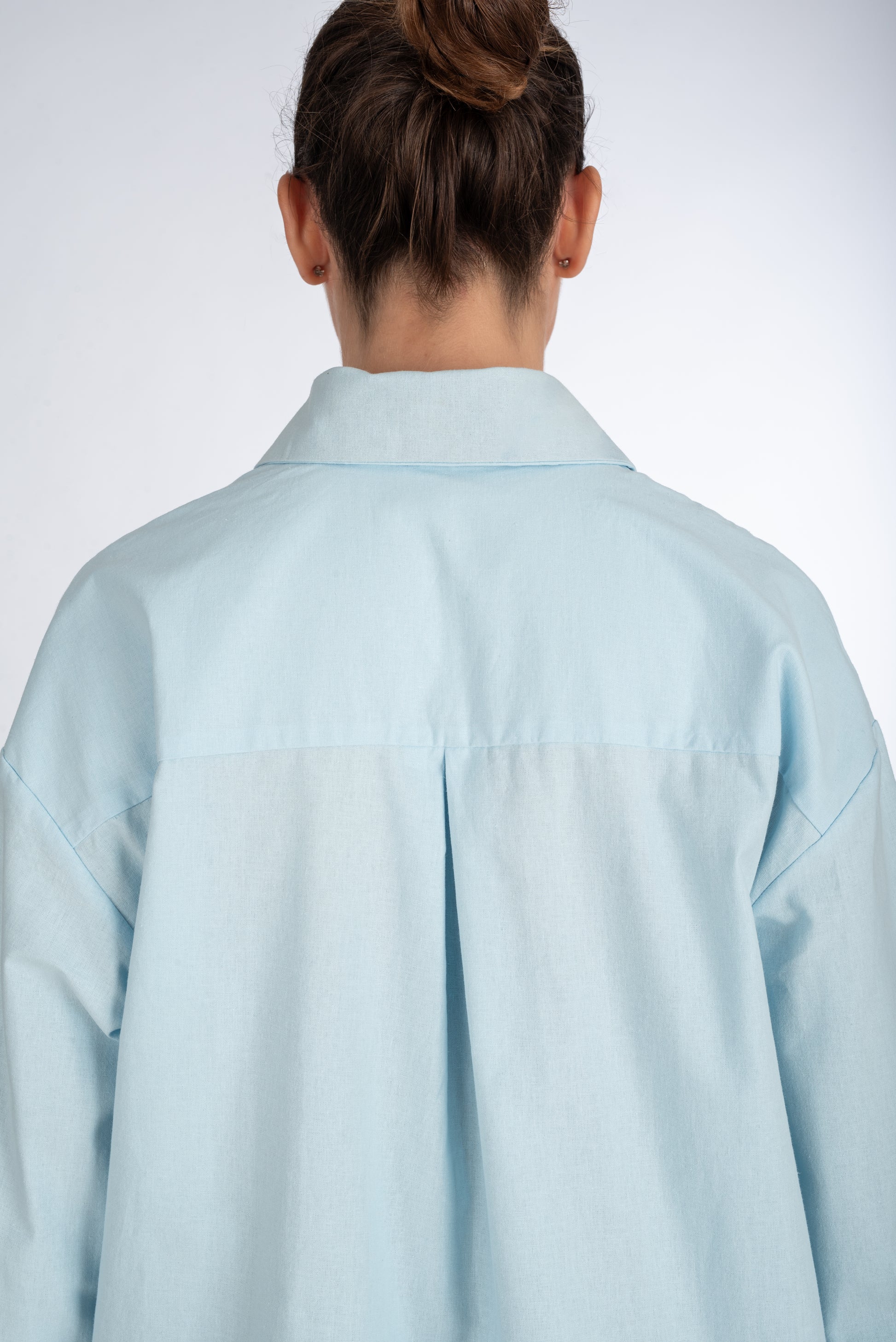 Lifestyle shot of the back of a baby blue women's shirt, highlighting the center pleat at the yoke and sleek, minimal design.